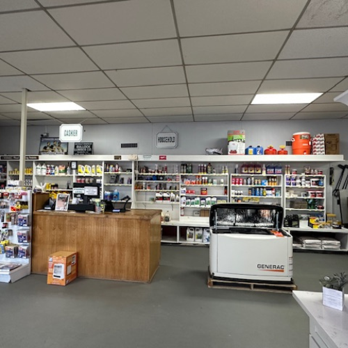 A hardware store interior with shelves stocked with various tools, paint cans, and supplies. A wooden cashier counter is in the center, with a large generator displayed nearby. The lighting is bright and even, creating a welcoming atmosphere.
