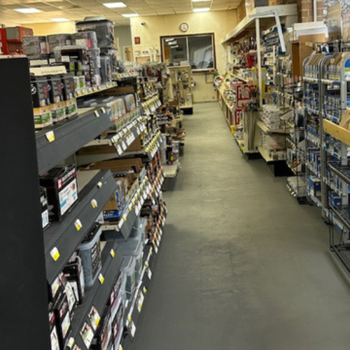 A hardware store aisle with shelves filled with various tools and supplies. The floor is gray, and fluorescent lights illuminate the space.