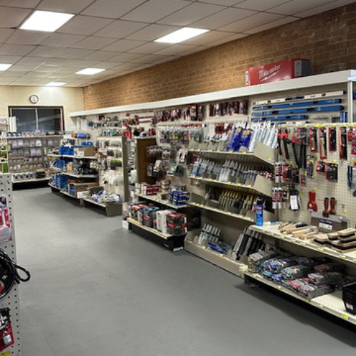A hardware store aisle with organized shelves displaying tools, paintbrushes, and supplies against a brick wall. Bright lighting creates a tidy, inviting atmosphere.
