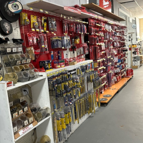 A hardware store aisle displays neatly arranged tools and supplies on shelves, with drill bits, saw blades, and packaged items. The store has a tidy, organized atmosphere.