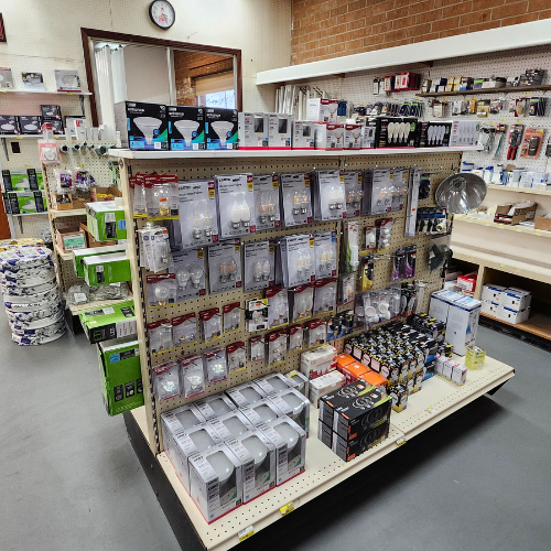A hardware store aisle displays various tools and accessories. The shelves are stocked with packaged light bulbs and batteries, creating an organized, utilitarian atmosphere.