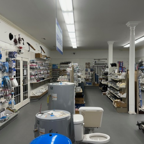 A well-lit hardware store aisle features shelves lined with plumbing supplies. Water heaters and a toilet display are central, suggesting a focus on home repairs.